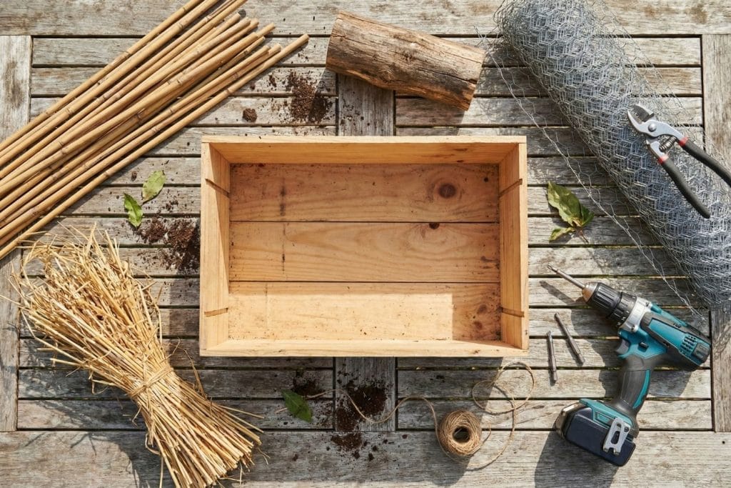 Matériel pour construire un hôtel à insectes : caisse à vin en bois, tiges de bambou, paille, perceuse et grillage sur une table de jardin